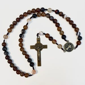 May include: A rosary with brown, black, and white agate beads. The rosary features a bronze crucifix with the inscription "INRI" and a circular medal. The beads are strung on a dark cord.