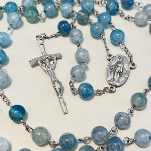 Op de afbeelding: Een rozenkrans met lichtblauwe en turquoise kralen. De rozenkrans heeft een zilverkleurig kruisbeeld en een zilverkleurige ovale hanger met een afbeelding van de Maagd Maria. De kralen zijn verbonden door een zilverkleurige ketting.