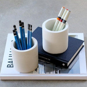 May include: Two white cylindrical pencil holders with pencils and pens on a white tray. One holder contains blue pencils, the other contains white pens with colored bands. The tray rests on a book with the word "BAUHAUS" visible.