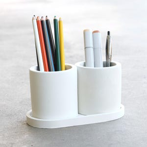 May include: A white desk organizer with two cylindrical compartments connected by a small, oval base. One compartment holds colored pencils, the other holds markers and a pen. The organizer is made of a smooth, matte material.