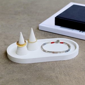 May include: A white, oval-shaped jewelry tray with two white cone ring holders and a bracelet. One ring is gold with a textured design, and the other is a gold beaded ring. The bracelet has silver and gray beads with a red cord.