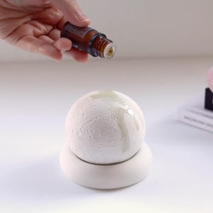 May include: A white ceramic essential oil diffuser with a textured, moon-like surface. A brown glass bottle is held above the diffuser, dispensing oil. A rose quartz face roller and books are in the background.