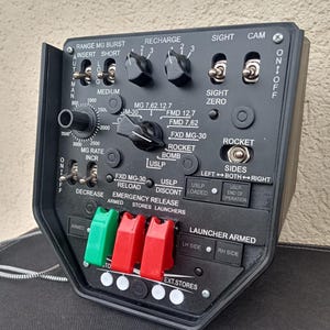 May include: A black control panel with various switches, dials, and labels. The panel includes text such as "RANGE MG BURST," "RECHARGE," and "ROCKET." It features green and red levers and is likely part of a military or industrial system.