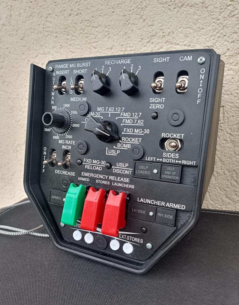 May include: A black control panel with various switches, dials, and labels. The panel includes text such as "RANGE MG BURST," "RECHARGE," and "ROCKET." It features green and red levers and is likely part of a military or industrial system.