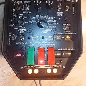 May include: A black control panel with numerous switches, dials, and indicator lights. The panel features labels such as "Range," "Recharge," and "Rocket." There are multiple toggle switches and rotary dials, along with illuminated buttons.