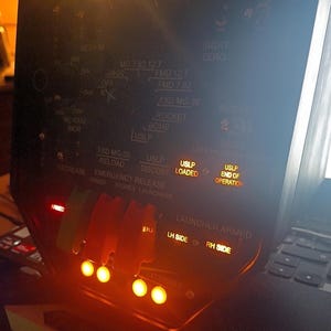 May include: A close-up of a black control panel with various switches, buttons, and illuminated indicators. The panel features text such as "LOADED", "ARMED", and "END OF OPERATION". The panel has a series of orange lights.