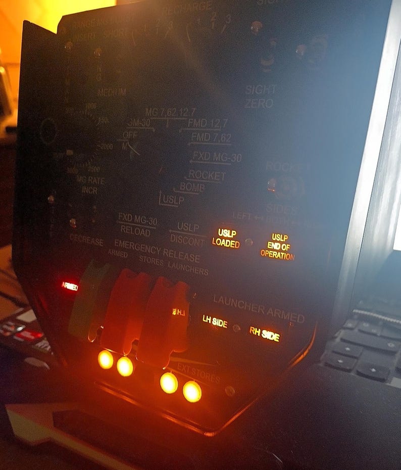 May include: A close-up of a black control panel with various switches, buttons, and illuminated indicators. The panel features text such as "LOADED", "ARMED", and "END OF OPERATION". The panel has a series of orange lights.