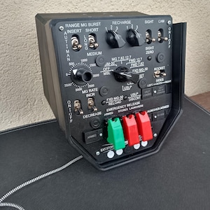 May include: A black control panel with various switches, dials, and labels. The panel includes text such as "RANGE MG BURST," "RECHARGE," and "ROCKET." It features multiple toggle switches and rotary dials, with green and red connectors at the bottom.