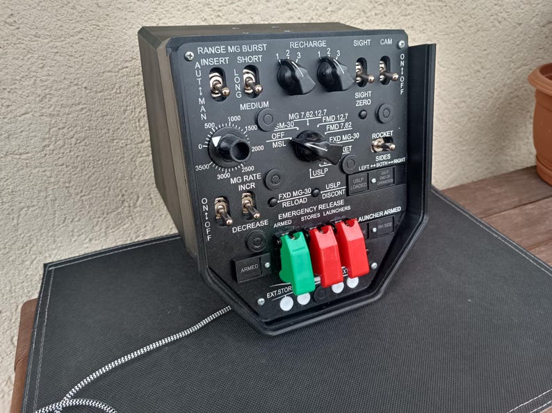 May include: A black control panel with various switches, dials, and labels. The panel includes text such as "RANGE MG BURST," "RECHARGE," and "ROCKET." It features multiple toggle switches and rotary dials, with green and red connectors at the bottom.