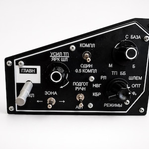 Puede incluir: Un panel de control negro con múltiples perillas, interruptores y etiquetas blancas. El panel tiene forma rectangular con una parte superior inclinada. El texto "ГЛАВН" es visible.