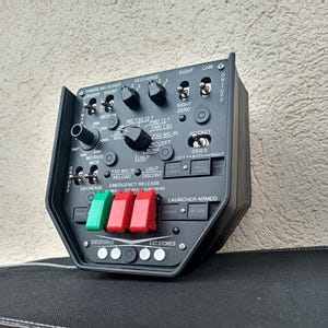 May include: A black control panel with various switches, dials, and buttons. The panel includes labels such as "Range MG Burst," "Recharge," and "Rocket." It features green, red, and white buttons labeled "Emergency Release" and "Ext. Stores."
