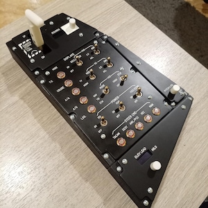 May include: Black control panel with numerous switches, buttons, and dials. The panel has white text and labels, including "ROUNDS", "MODE", and "CRUISE". It features a white lever and a digital display. The panel is likely for a flight simulator or aircraft.