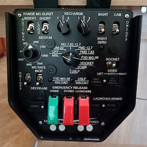 May include: A black control panel with various switches, dials, and labels. Features include "RANGE MG BURST," "RECHARGE," and "ROCKET." It has green and red levers labeled "ARMED" and "STORES."