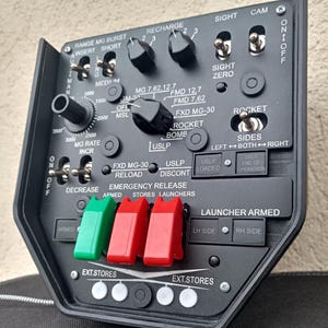 May include: A black control panel with various switches, dials, and buttons. The panel includes labels such as "Range MG Burst," "Recharge," and "Rocket." It features red and green toggle switches and a series of white and black buttons.