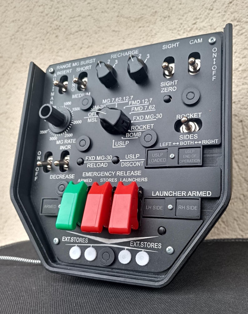 May include: A black control panel with various switches, dials, and buttons. The panel includes labels such as "Range MG Burst," "Recharge," and "Rocket." It features red and green toggle switches and a series of white and black buttons.