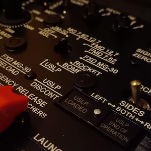 May include: Close-up of a black control panel with white lettering and various switches and buttons. Text includes "USLP", "ROCKET", and "LOADED". A red switch is visible on the left.