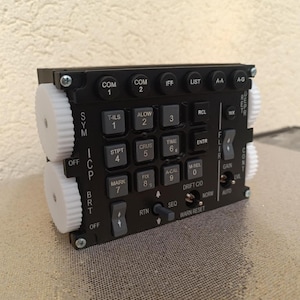 Puede incluir: Un panel de control negro con numerosos botones y diales, con texto y etiquetas blancos. El panel incluye botones etiquetados como "COM", "IFF", "LIST" y "ENTR", junto con diales giratorios en los lados. Diseñado para aviación o uso técnico.