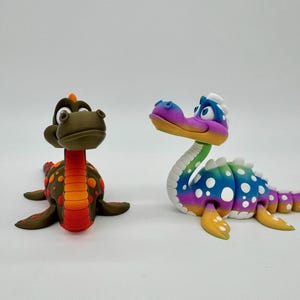 Könnte beinhalten: Zwei handgefertigte Dinosaurierfiguren. Eine ist braun mit orangefarbenen und roten Akzenten. Die andere ist eine lebendige Mischung aus Blau, Lila, Grün und Gelb mit weißen Punkten. Beide liegen.