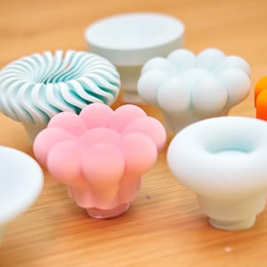 May include: A collection of colorful, decorative knobs in various shapes and designs. The knobs include a light blue swirled design, a pink flower shape, and a white rounded shape. Other knobs are white, orange, and light blue.