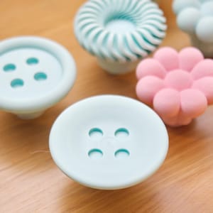 Peut inclure: Une collection de boutons décoratifs de différentes formes et couleurs. Les boutons comprennent un bouton bleu clair, une forme de fleur rose et d'autres formes uniques. Ces boutons sont probablement destinés aux meubles ou à la décoration.