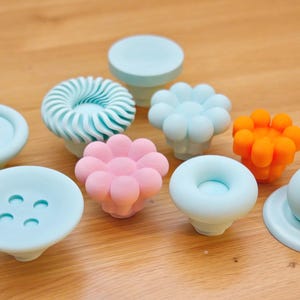 Może przedstawiać: Kolekcja dekoracyjnych gałek do szafek w różnych kształtach i kolorach. Gałki obejmują jasnoniebieskie, różowe i pomarańczowe wzory, takie jak kształty kwiatów i guzików. Ułożone są na drewnianej powierzchni.