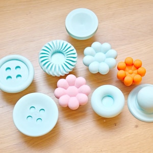 Könnte beinhalten: Eine Sammlung farbenfroher Silikonknöpfe und -griffe in verschiedenen Formen, darunter Knöpfe, Blumen und ein Donut. Die Farbpalette umfasst Hellblau, Rosa und Orange. Diese dekorativen Beschlagteile sind auf einer hellen Holzoberfläche angeordnet.