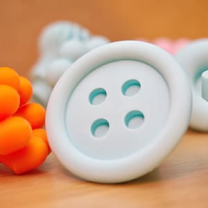 May include: A large, light blue button with four holes, set against a blurred background of other craft supplies. An orange, textured object is in the foreground. The button is on a wooden surface.