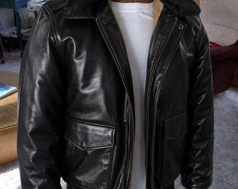 May include: A black leather bomber jacket featuring a dark faux fur collar, a front zip fastening, two flap pockets at the waist, and ribbed cuffs. The jacket is displayed on a mannequin wearing a plain white t-shirt underneath the open front.