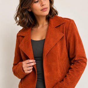 Könnte beinhalten: Eine rostorangefarbene Wildlederjacke mit breitem Reverskragen, langen Ärmeln und geradem Saum. Die Jacke wird über einem schlichten dunkelgrauen Tanktop getragen. Die Textur des Stoffes ist matt und gleichmäßig, was für ein strukturiertes Aussehen sorgt.