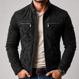 Puede incluir: Una chaqueta de ante negro carbón con cuello alzado, cremallera frontal completa y dos bolsillos horizontales con cremallera en el pecho. La chaqueta se lleva sobre una camiseta lisa de color beige claro, combinada con vaqueros azules y un cinturón de cuero marrón.