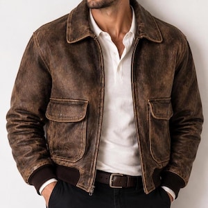 Könnte beinhalten: Eine braune Leder-Bomberjacke in Distressed-Optik mit klassischem Kragen, Frontreißverschluss und zwei großen Pattentaschen auf der Vorderseite. Die Jacke verfügt über dunkelbraune Rippbündchen an den Ärmeln und am Saum für einen Sitz.