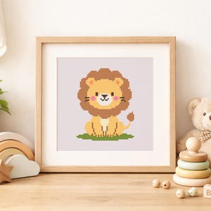 May include: A square wooden frame containing a cross-stitch pattern of a cartoon lion. The lion has a brown mane, yellow body, pink cheeks, and sits on a patch of green grass against a light purple grid background, centered within the frame.