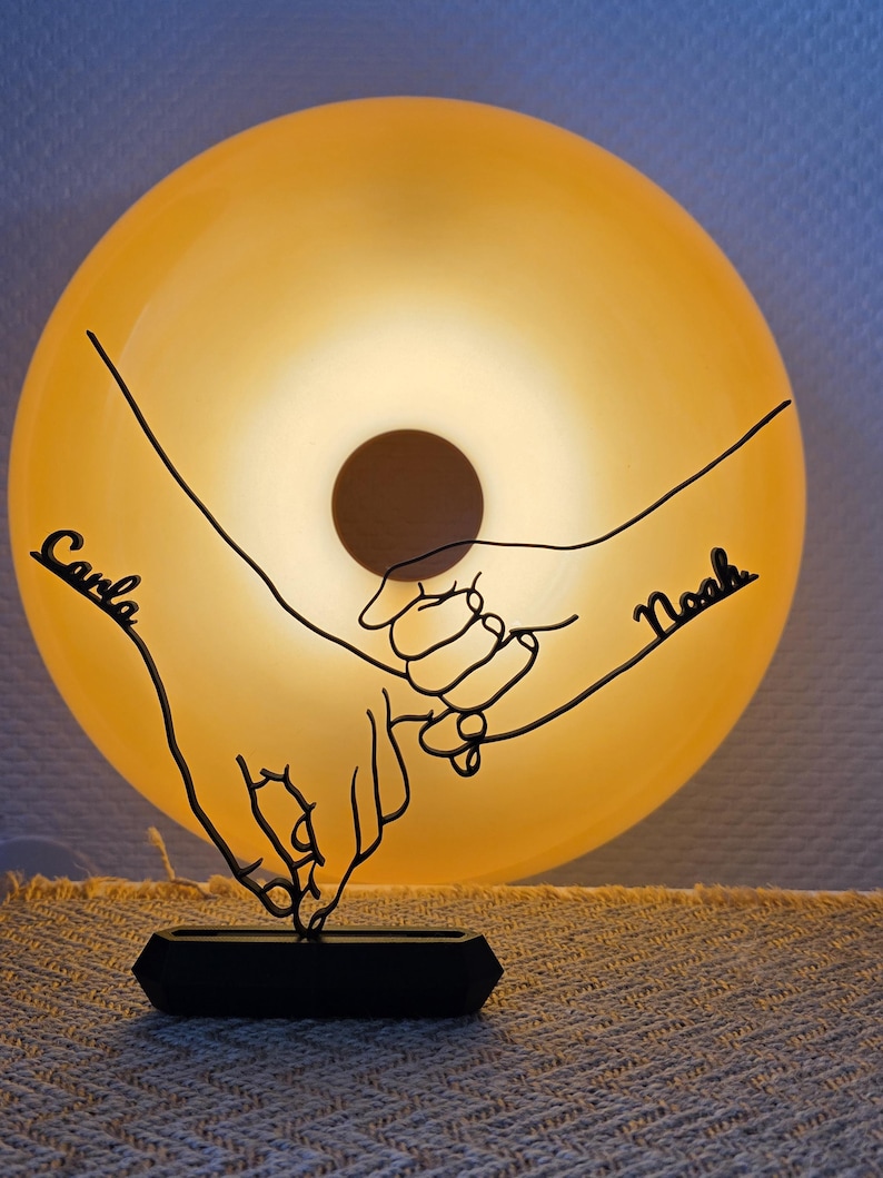 May include: A black metal sculpture of two hands holding each other, with the names "Carla" and "Noah" inscribed on each hand. The sculpture is set against a glowing, round, yellow light. The base is a black, rectangular shape.