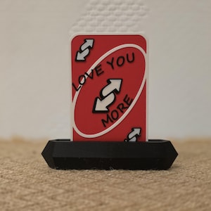 May include: A red and white "Love You More" Uno card in a black holder. The card has white arrows and text on a red background. The holder is a geometric shape.