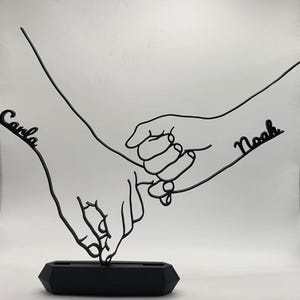 May include: Black metal sculpture depicting two hands holding each other. The names "Carla" and "Noah" are written on the sculpture. The sculpture is mounted on a black, geometric base. The artwork is a symbol of connection and love.