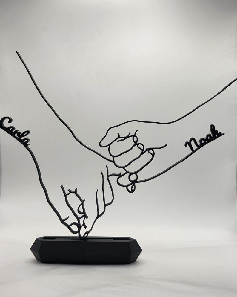 May include: Black metal sculpture depicting two hands holding each other. The names "Carla" and "Noah" are written on the sculpture. The sculpture is mounted on a black, geometric base. The artwork is a symbol of connection and love.