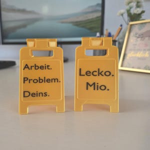 Könnte beinhalten: Zwei gelbe Schilder mit schwarzem Text. Auf dem linken Schild steht "Arbeit. Problem. Deins." Auf dem rechten Schild steht "Lecko. Mio.". Die Schilder stehen auf einer weißen Oberfläche.