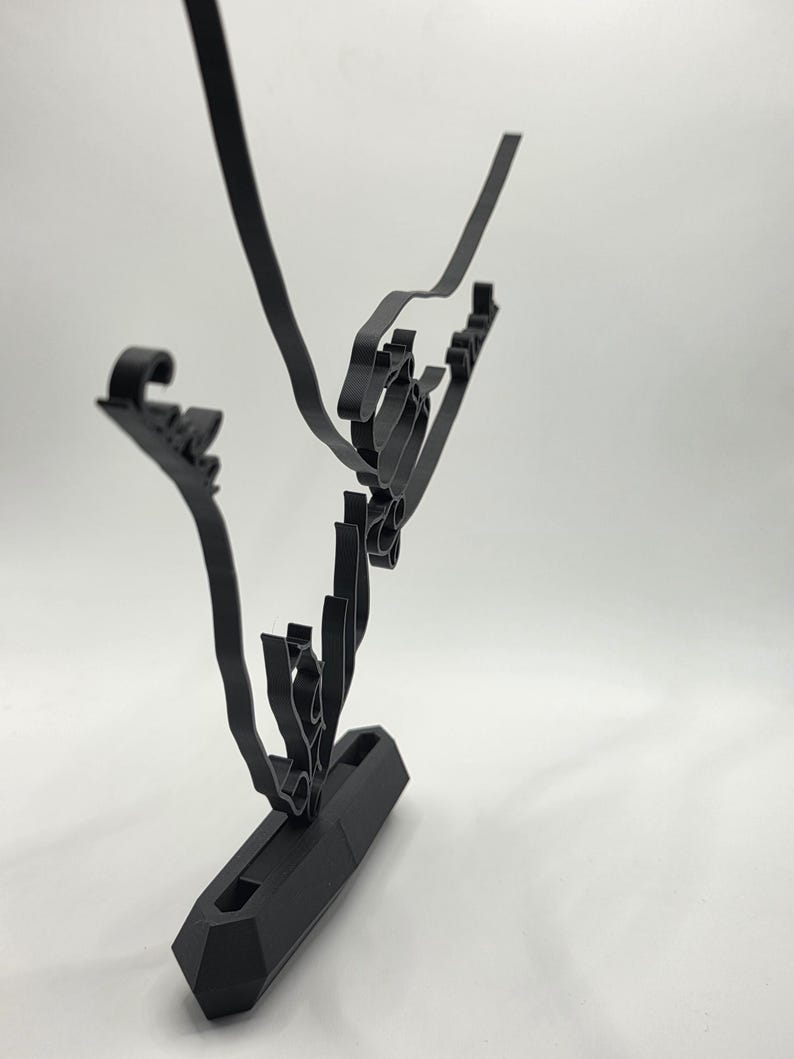 May include: A black, abstract metal sculpture with a geometric base and flowing, linear elements. The sculpture has a modern design, with curved and angular shapes. The piece is set against a white background, highlighting its form.