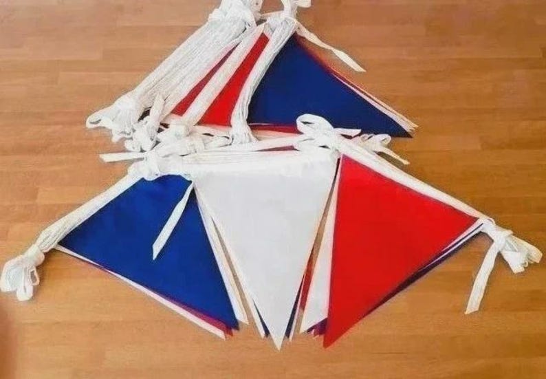 May include: A set of triangular bunting flags in red, white, and blue. The flags are attached to a white fabric cord with ties at the ends. The pennants are arranged in overlapping layers, showcasing a repeating pattern of solid primary colors.
