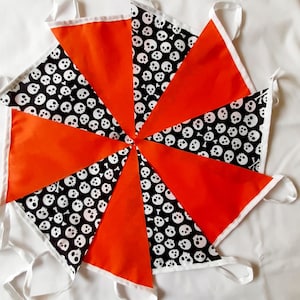 May include: A Halloween-themed bunting with alternating orange and black skull-patterned pennants. Each triangular flag is trimmed with white fabric and has a white ribbon for hanging. The bunting is arranged in a circular pattern.