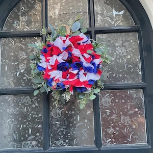 May include: A patriotic wreath with red, white, and blue paper flowers, accented with green foliage. The wreath is displayed on a window with a floral pattern.