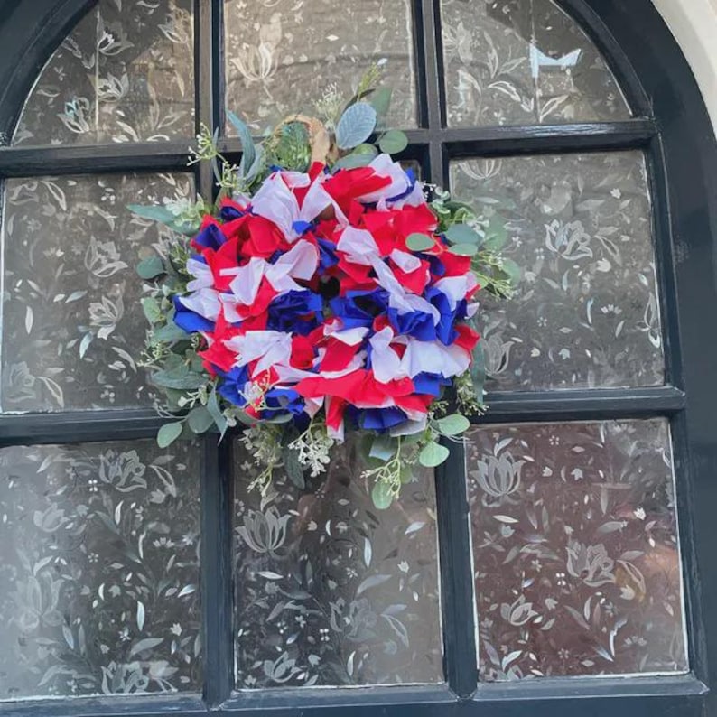 May include: A patriotic wreath with red, white, and blue paper flowers, accented with green foliage. The wreath is displayed on a window with a floral pattern.