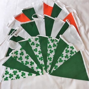 May include: A selection of triangular fabric bunting flags in green, white, and orange. Some flags have a shamrock pattern. The flags are arranged in a fan shape on a white surface.