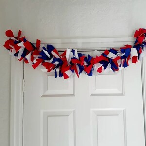 May include: A festive garland made of red, white, and blue fabric strips, draped above a white door. The fabric is knotted and arranged to create a decorative accent, perfect for patriotic holidays or themed events.
