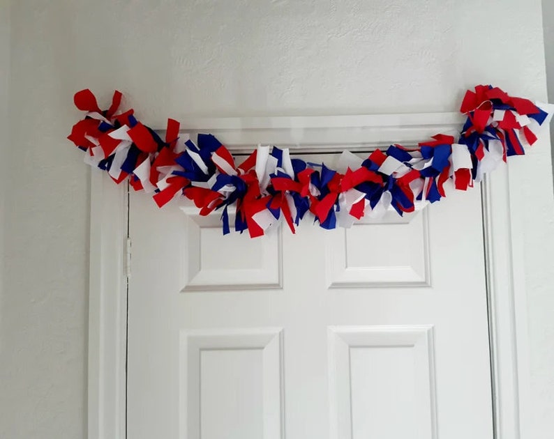 May include: A festive garland made of red, white, and blue fabric strips, draped above a white door. The fabric is knotted and arranged to create a decorative accent, perfect for patriotic holidays or themed events.