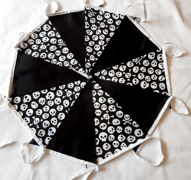 May include: A black and white skull and crossbones banner. The banner is made of triangular fabric pieces, alternating between solid black and a white skull pattern. White trim and ties complete the design.
