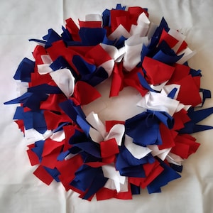 May include: A handmade wreath composed of red, white, and blue fabric strips. The wreath is circular, with the fabric strips arranged to create a textured, layered effect. The colors are reminiscent of the American flag.