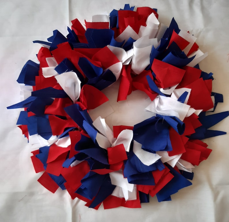 May include: A handmade wreath composed of red, white, and blue fabric strips. The wreath is circular, with the fabric strips arranged to create a textured, layered effect. The colors are reminiscent of the American flag.