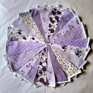 May include: A circular arrangement of triangular fabric bunting flags in various shades of purple and white. The flags feature diverse patterns including floral prints, gingham checks, polka dots, and solid light purple fabric.