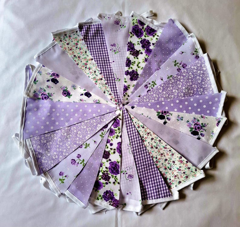May include: A circular arrangement of triangular fabric bunting flags in various shades of purple and white. The flags feature diverse patterns including floral prints, gingham checks, polka dots, and solid light purple fabric.